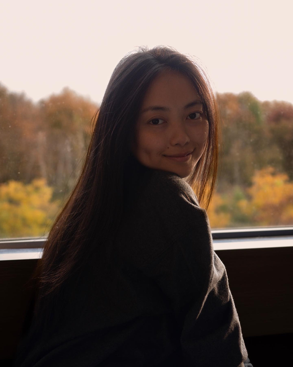 Mai Vee Vang sitting in front of a window with the view of golden tree foilllage, smiling back endearingly at the camera.  